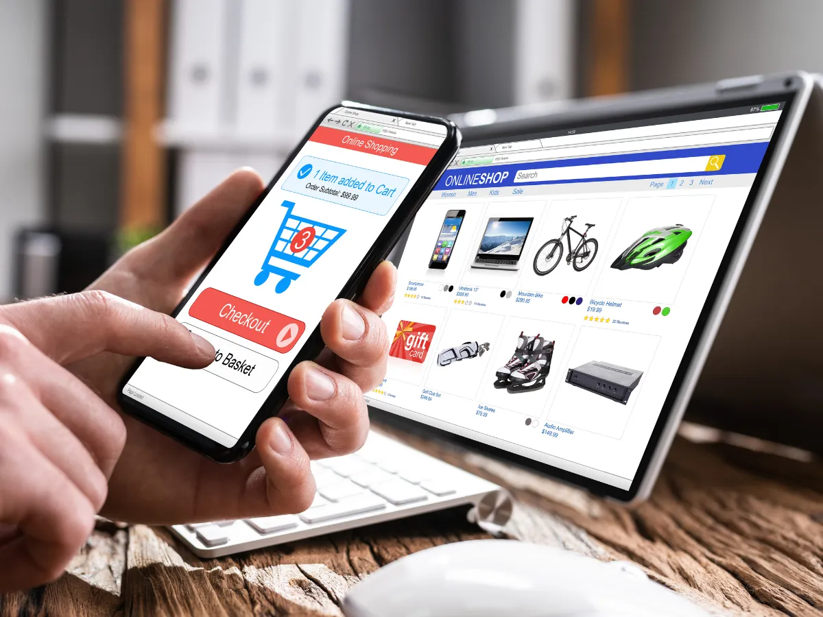 A hand holding a phone showing an online shopping cart, with a laptop store page behind it
