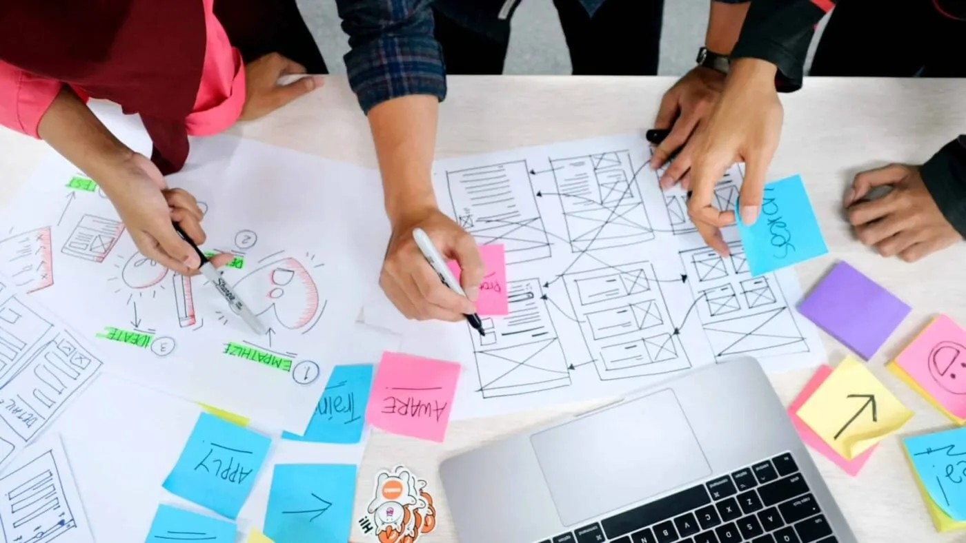 Hands sketching website wireframes and sticky notes on a table beside a laptop